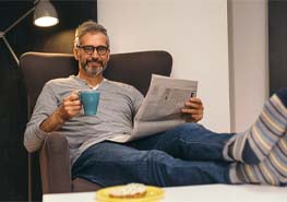 a person sitting in a chair, reading the newspaper, and enjoying a piece of toast and cup of coffee